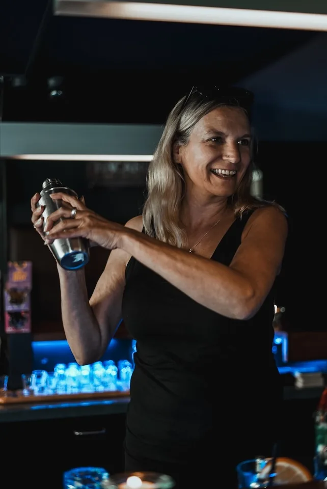 Smiling bartender in black top shaking a cocktail shaker behind the bar with blue lighting.