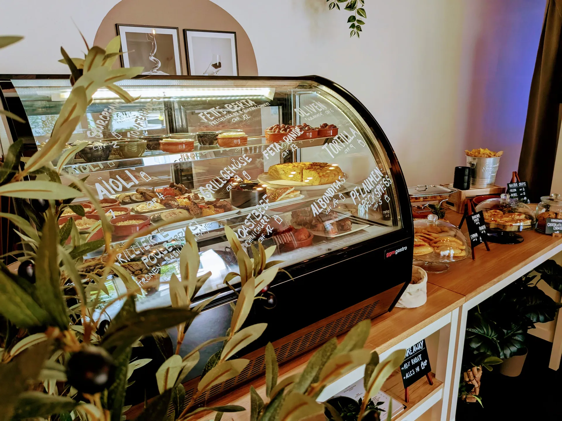 Curved glass deli display counter filled with Spanish tapas, tortilla, olives, and labeled small dishes.