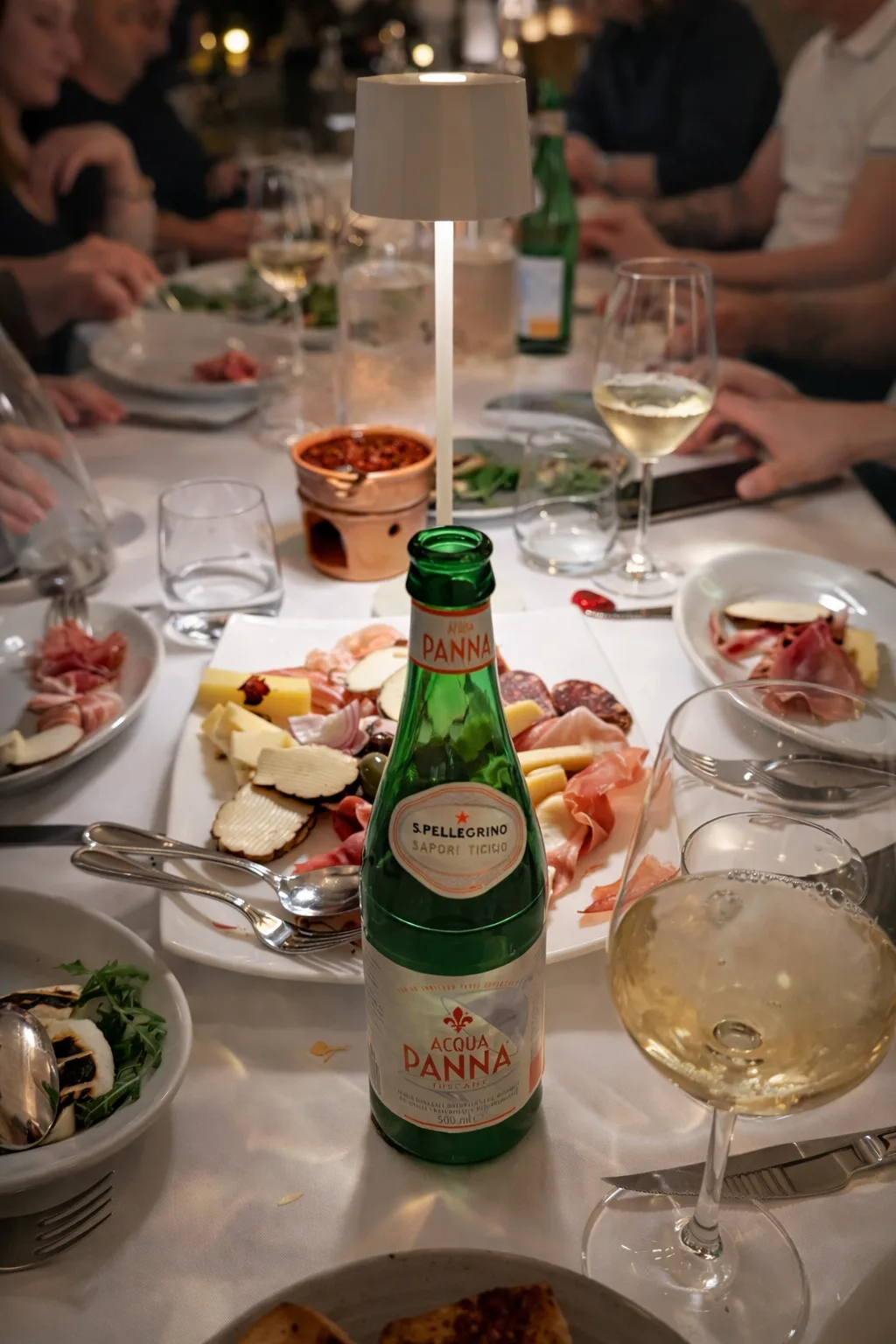 Lively dinner table spread with charcuterie, salads, white wine, and Acqua Panna sparkling water.