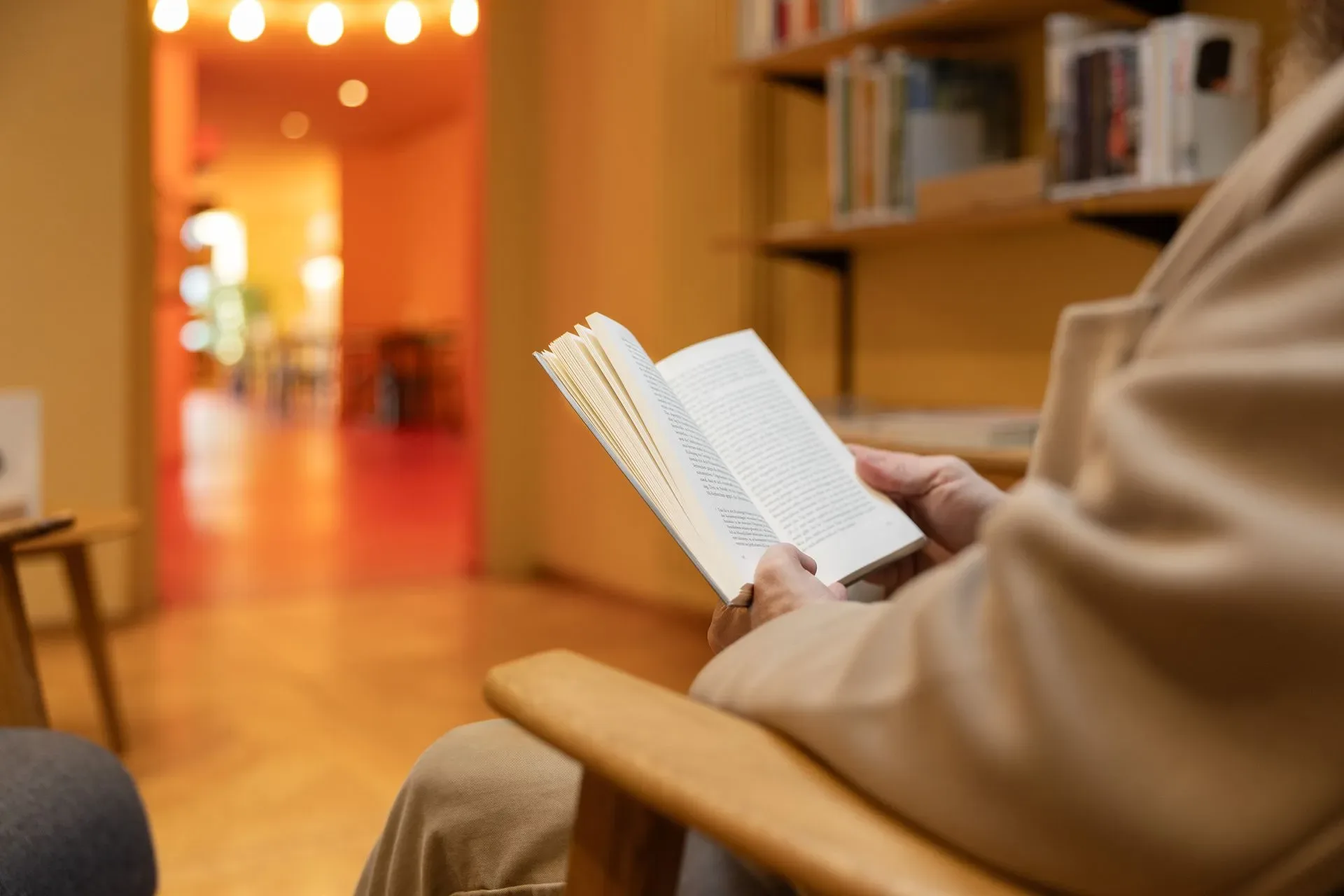 A guest in a beige coat reads a book in the warm-toned café library area.