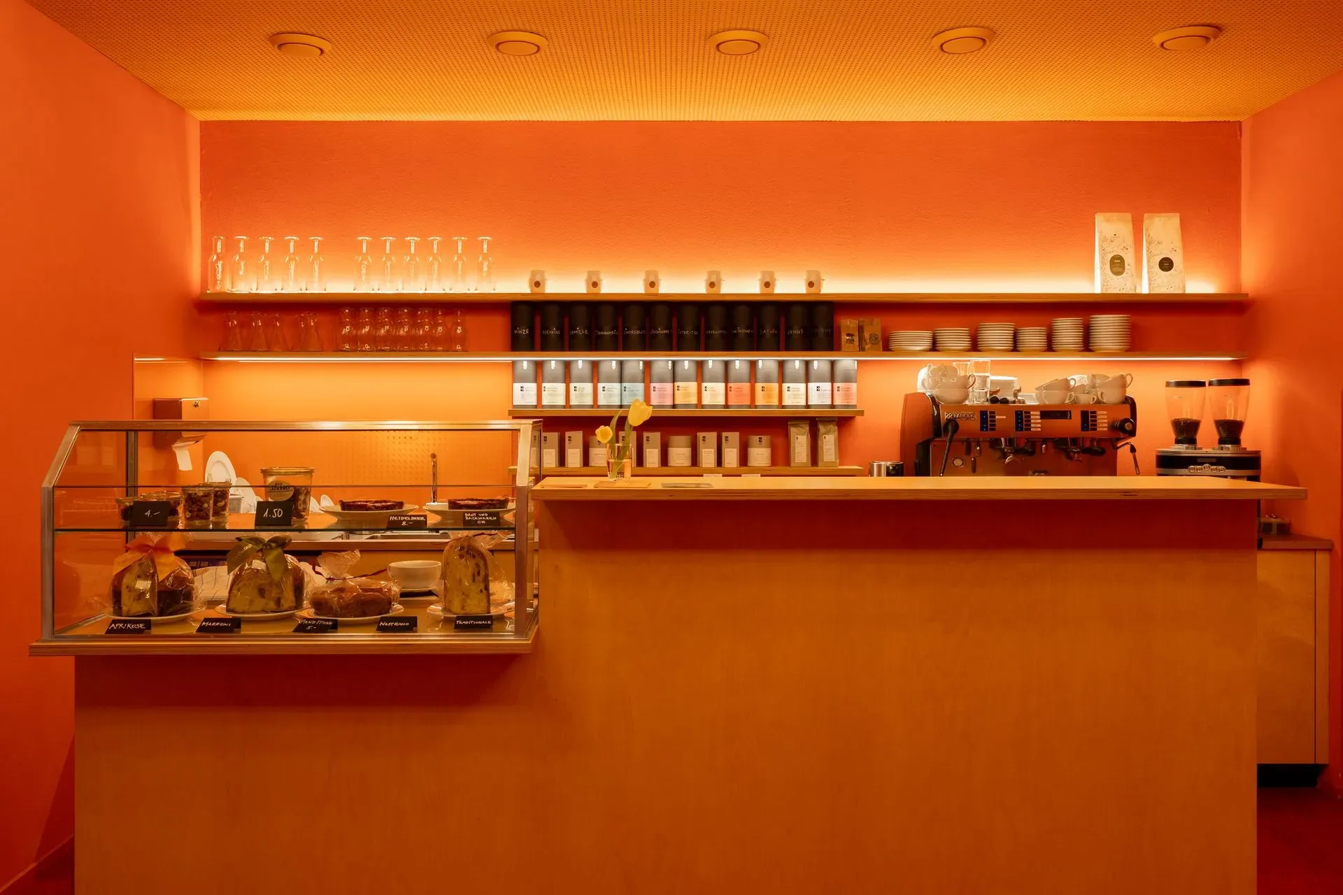 The café counter displaying pastries, an espresso machine, and backlit shelves of tea and glassware.