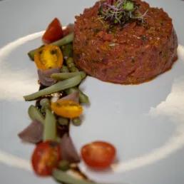 Elegant beef tartare moulded with green beans and cherry tomatoes on a white plate.