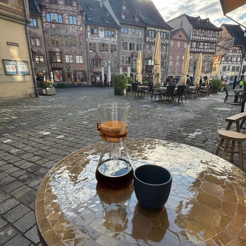 A Chemex pour-over and ceramic cup on a mosaic table in a charming European old-town square.