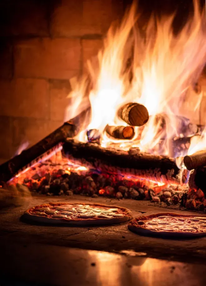 Two small pizzas baking on the stone floor of a roaring wood-fired oven.