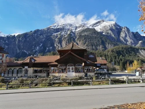 A grand alpine restaurant building set against dramatic snow-capped mountains under a blue sky.