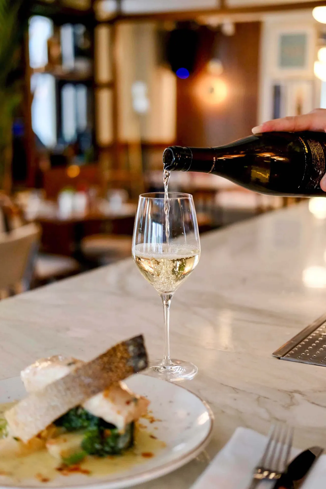 White wine being poured into a tall glass beside a plated fish dish at a marble bar counter.