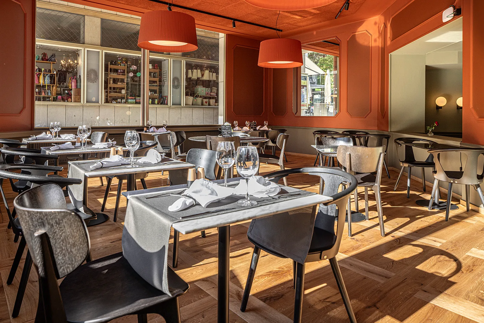 Sunlit terracotta-red dining room with set tables, wine glasses, and large pendant lamps creating a welcoming atmosphere.