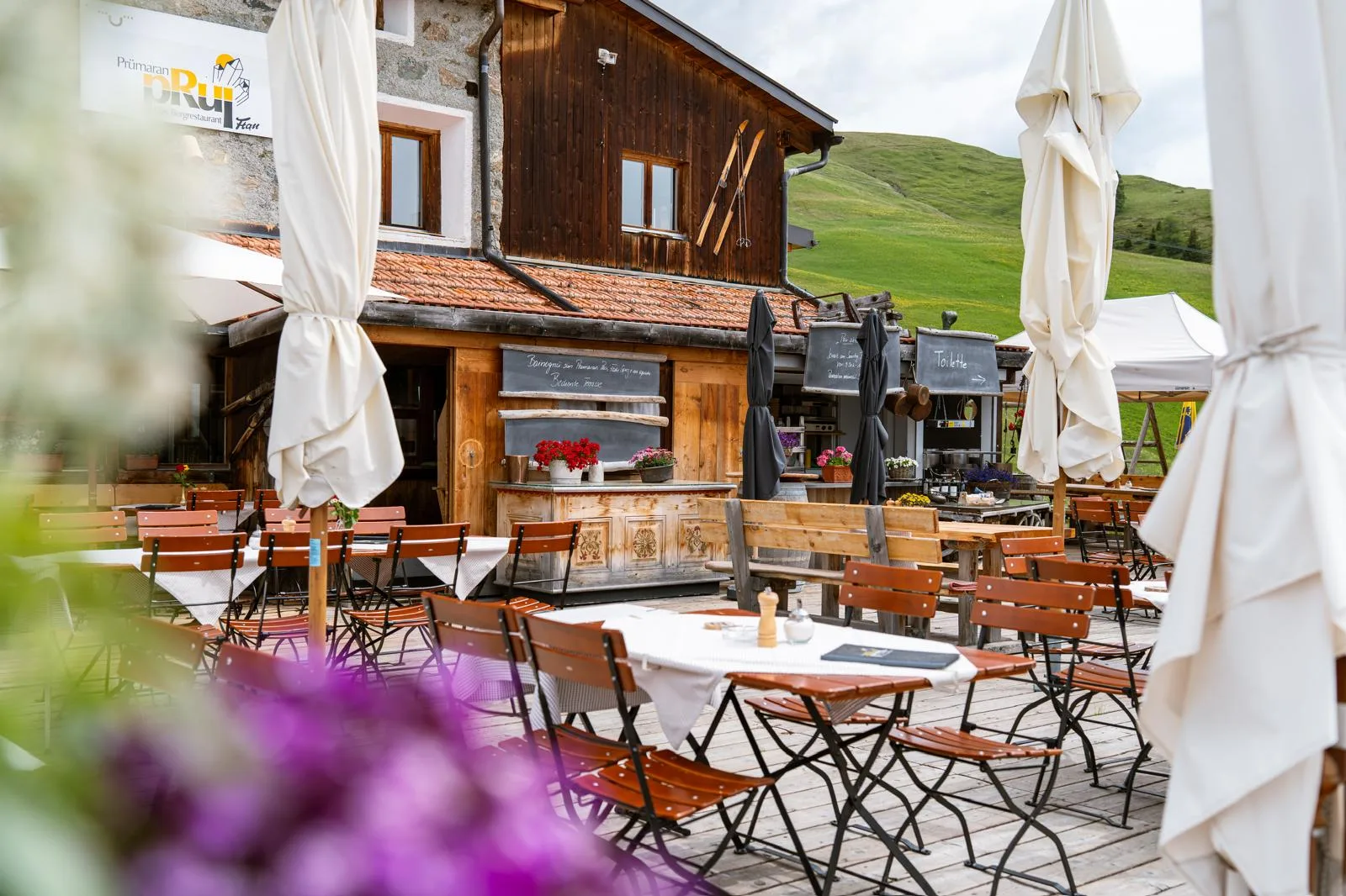 Sunny alpine terrace of Prümaran Prui restaurant with wooden chairs and green mountain backdrop.