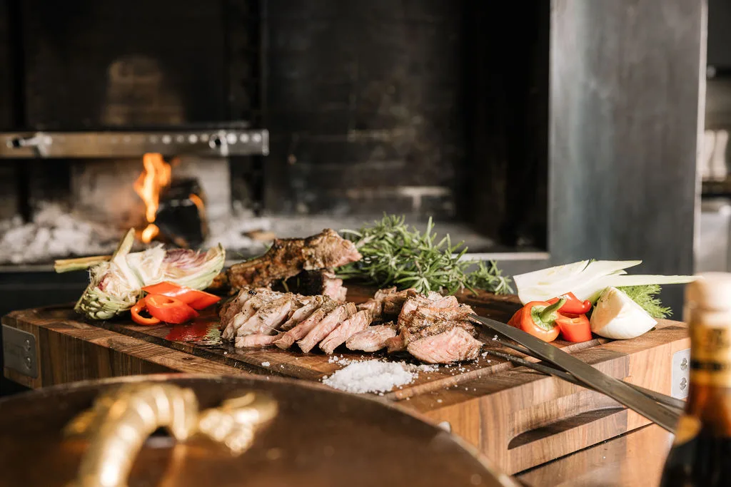 Sliced grilled meat on a wooden board with rosemary and vegetables beside an open wood fire.