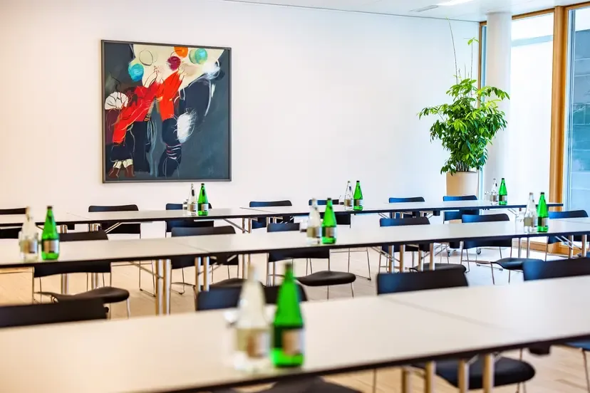 Modern seminar room with rows of tables, water bottles, black chairs, and a colorful abstract artwork on the wall.
